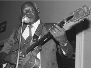 Johnnie Billington standing playing the guitar and talking into a microphone.